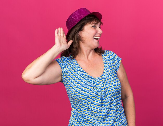 Divertente donna di mezza età con cappello da festa che guarda da parte con la mano sull'orecchio sorridendo allegramente