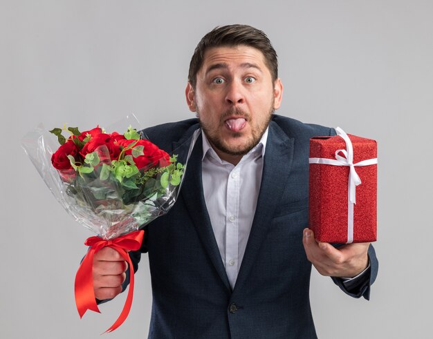 Divertente e gioioso giovane bell'uomo che indossa un abito con un mazzo di rose e un regalo per san valentino che tira fuori la lingua