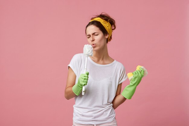 Divertente giovane casalinga europea che indossa la fascia, t-shirt casual e guanti di gomma verdi che si godono il processo di pulizia a casa, tenendo la spugna e lo scopino alla bocca come un microfono e firmando