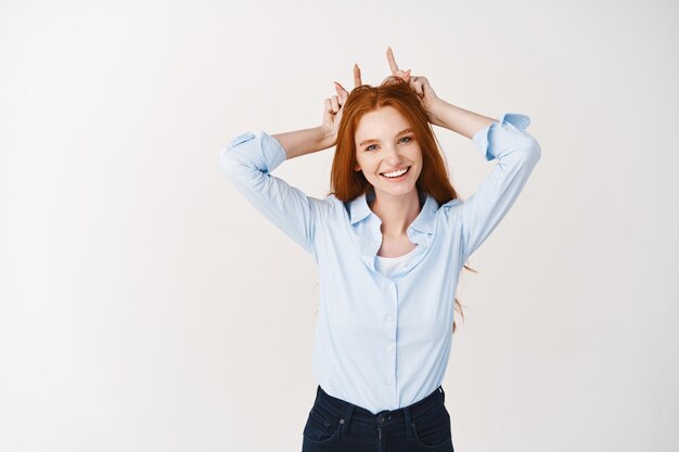 Divertente giovane donna con i capelli rossi e le lentiggini scherzare, mostrando corna da diavolo con le dita sulla testa e sorridente, in piedi sul muro bianco