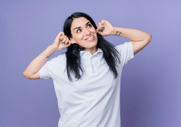 Divertente giovane ragazza caucasica bruna tira fuori la lingua e tiene le orecchie isolate sulla parete viola