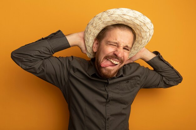 Divertente giovane uomo bello in camicia grigia e cappello estivo che fa smorfia tenendosi per mano dietro il collo