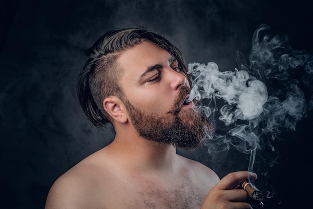 Divertente maschio barbuto senza camicia che fuma un sigaro su sfondo grigio.