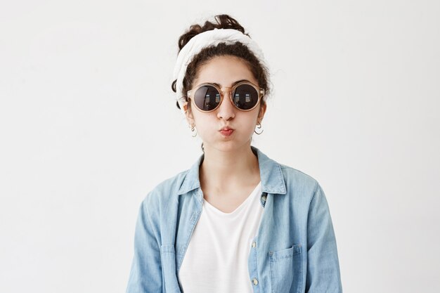 Divertente modello femminile con capelli scuri in labbra da broncio, camicia di jeans e occhiali da sole rotondi, fa una smorfia come stand. La bella donna allegra si diverte al chiuso, soffia le guance