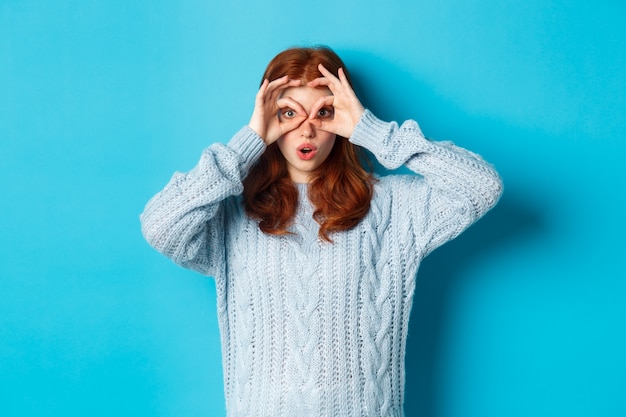 Divertente modello femminile rossa in maglione, fissando la telecamera attraverso gli occhiali delle dita, vedendo qualcosa di interessante, in piedi su sfondo blu