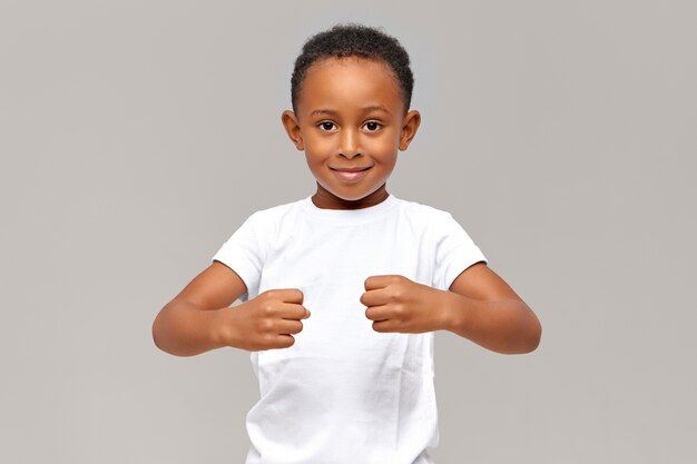 Divertente ragazzo africano di dieci anni in maglietta bianca tenendo i pugni chiusi davanti a sé dimostrando forza o tenendo oggetti invisibili
