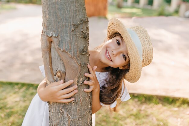 Divertente ragazzo dai capelli scuri con grandi occhi e sorriso che abbraccia albero nel parco. Ritratto all'aperto della bambina felice in cappello di paglia che gode delle vacanze estive.