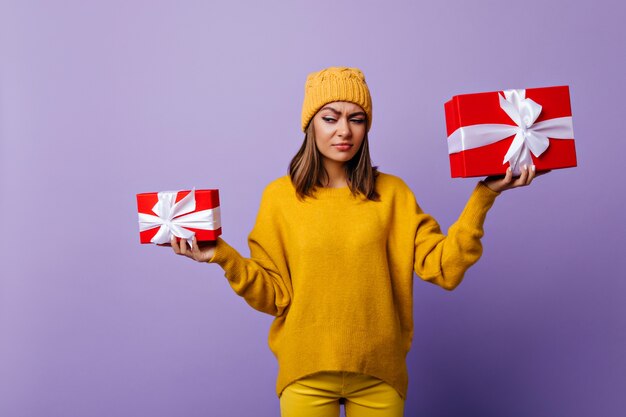 Divertente signora dai capelli scuri in cappello alla moda che tiene le caselle presenti. Ritratto di attraente donna bruna in maglione giallo in posa prima di Natale.