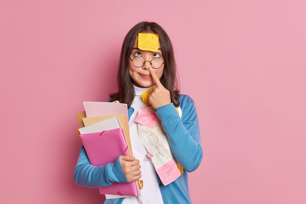 Divertente studente nerd tocca il naso ha una nota adesiva con grafica attaccata sulla fronte tiene cartelle e documenti concentrati sopra si prepara per la sessione di esame. La donna studia documenti con somme in ufficio.