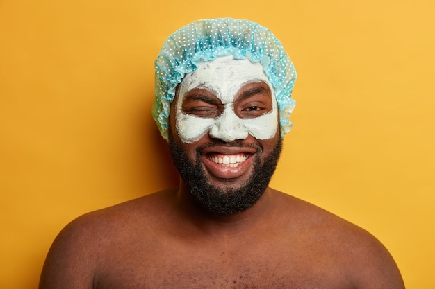 Divertente uomo dalla pelle scura positiva ammicca gli occhi, applica una maschera di argilla facciale antiaging dopo aver fatto la doccia, indossa un copricapo protettivo