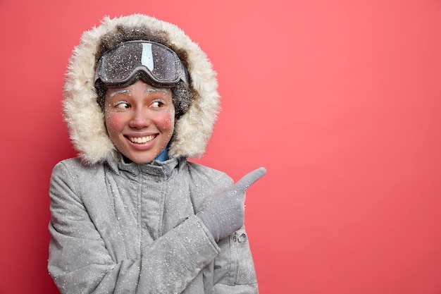 Divertimento invernale e concetto di sci. La donna afroamericana dalla pelle scura soddisfatta in abbigliamento esterno indica la direzione verso il luogo di villeggiatura per godersi la stagione di dicembre.