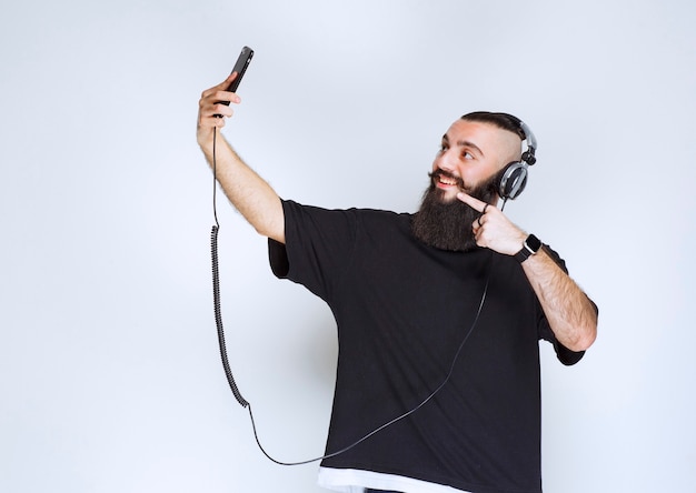 Dj con la barba che indossa le cuffie aprendo la fotocamera dello smartphone e prendendo il suo selfie.