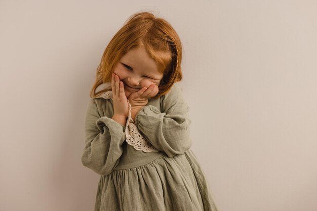 Dolce bambina dalla pelle chiara con capelli rossi in abiti casual tocca le sue guance su sfondo bianco Concetto di bambini