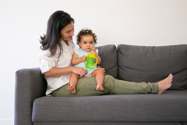 Dolce bambina seduta sulle ginocchia di mamme e acqua potabile dalla bottiglia. Copia spazio. Genitorialità e concetto di infanzia