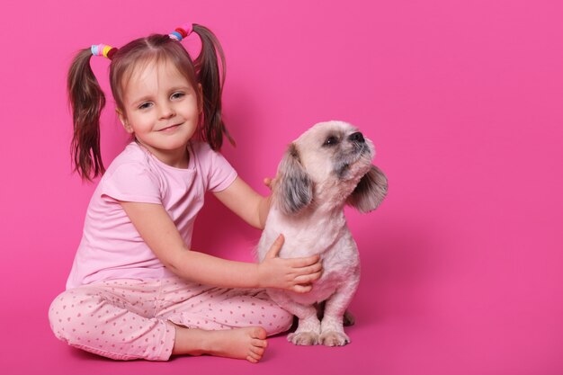 dolce piccolo bambino carino con due code di cavallo divertenti, essendo di buon umore, trascorrendo il tempo libero con l'animale domestico, tenendolo vicino, cucciolo guardando da parte, odorando, seduto calmo. Copyspace per la pubblicità.