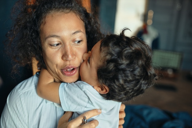 Dolce ritratto di tenero ragazzino di razza mista che bacia giovane la sua mamma eccitata sulla guancia, tenendo le braccia intorno al collo