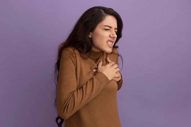 Dolorante con gli occhi chiusi giovane bella ragazza che indossa un maglione a collo alto marrone che mette la mano sul cuore isolato sul muro viola con spazio di copia