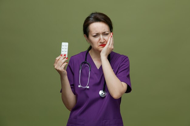 Dolorante dottoressa di mezza età che indossa uniforme e stetoscopio intorno al collo che mostra un pacchetto di compresse tenendo la mano sulla guancia con gli occhi chiusi che hanno mal di denti isolati su sfondo verde oliva