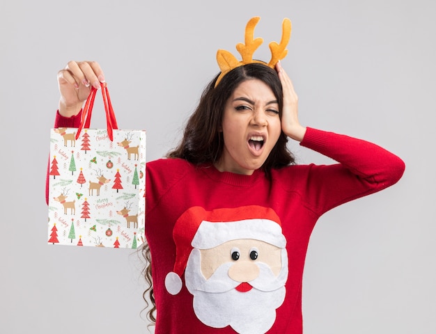 Dolorante giovane bella ragazza che indossa la fascia di corna di renna e maglione di Babbo Natale che tiene il sacchetto del regalo di Natale guardando la telecamera tenendo la mano sulla testa avendo mal di testa isolato su sfondo bianco