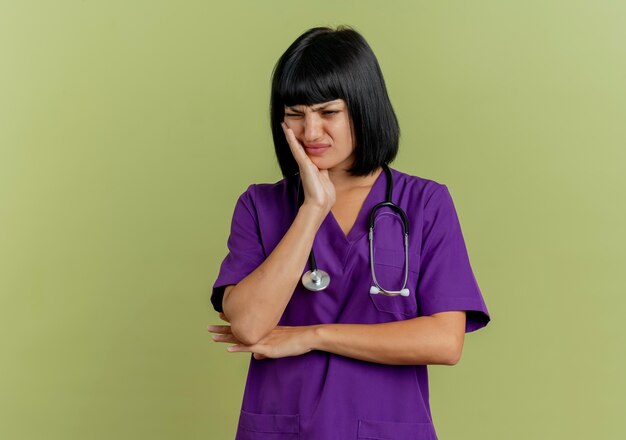 Dolorante giovane donna bruna medico in uniforme con lo stetoscopio mette la mano sul viso isolato su sfondo verde oliva con lo spazio della copia