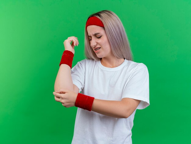 Dolorante giovane donna sportiva con le parentesi graffe che indossa la fascia e braccialetti mette la mano sul gomito isolato sulla parete verde