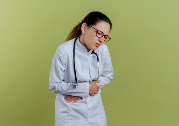 Dolorante giovane medico femminile che indossa abito medico e stetoscopio con gli occhiali ha afferrato lo stomaco isolato sulla parete verde oliva