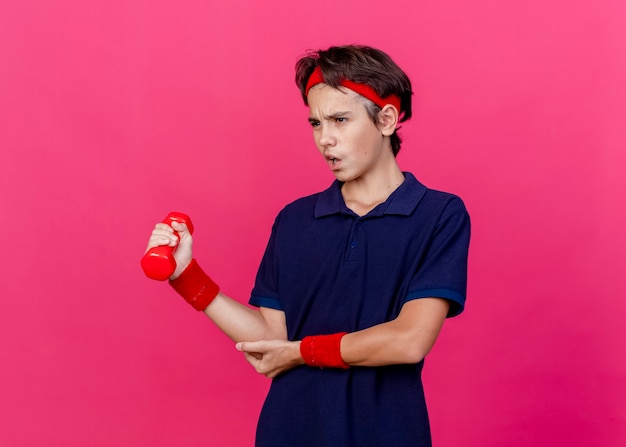 Dolorante giovane ragazzo sportivo bello che indossa la fascia e braccialetti con bretelle dentali guardando dritto tenendo il manubrio mettendo la mano sul gomito isolato su sfondo cremisi con lo spazio della copia