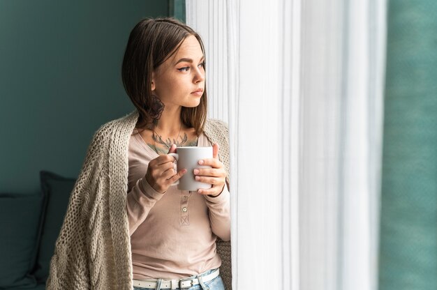 Donna a casa prendendo un caffè e guardando attraverso la finestra durante la pandemia