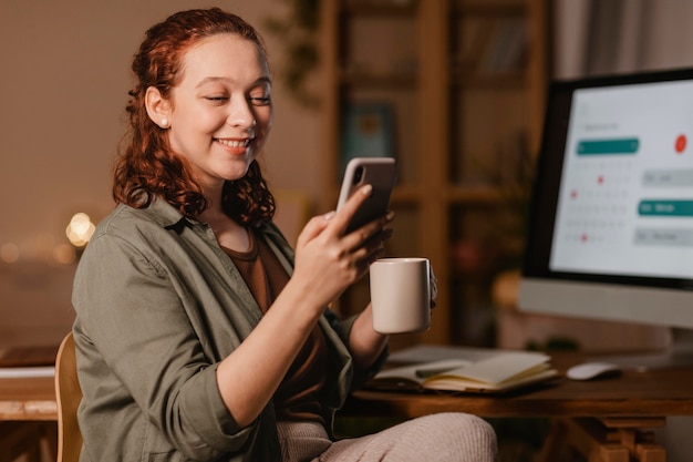 Donna a casa utilizzando smartphone davanti al computer mentre beve il caffè