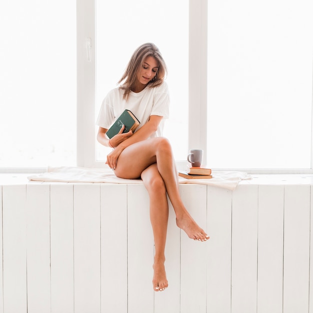 Donna a piedi nudi con il libro seduto sul davanzale della finestra