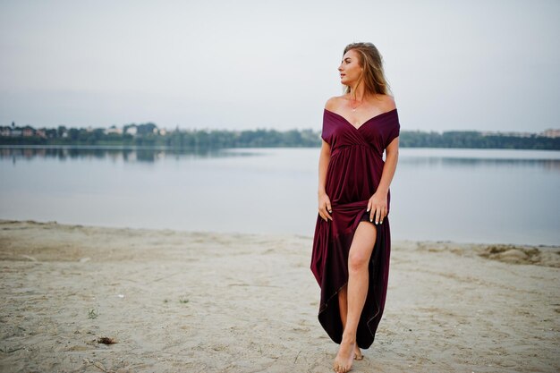 Donna a piedi nudi sensuale bionda in vestito rosso da marsala che posa contro il lago sulla sabbia