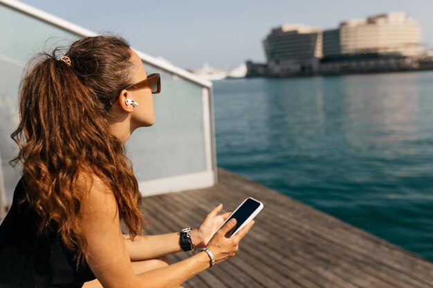 Donna abbastanza affascinante con capelli lunghi ondulati che indossa un top nero e occhiali da sole in cuffie wireless che tengono smartphone e guardano avanti o al lago alla luce del sole