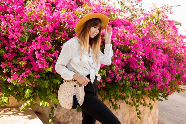 Donna abbastanza bionda in cappello di paglia che posa sopra l'albero di fioritura rosa nel giorno di molla soleggiato