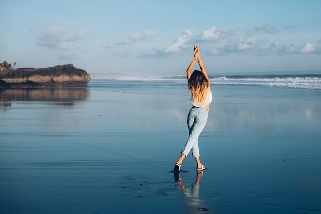 Donna abbastanza caucasica adatta in top bianco e jeans sulla spiaggia riflettente dall'oceano alla luce del tramonto