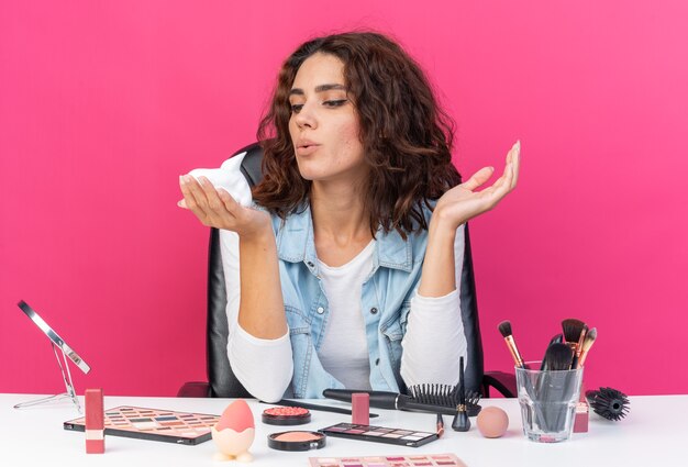 Donna abbastanza caucasica contenta seduta al tavolo con strumenti per il trucco che tiene e guarda la mousse per capelli isolata sulla parete rosa con spazio per le copie