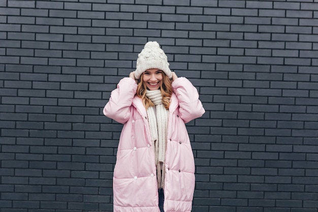 Donna abbastanza caucasica in abbigliamento invernale in posa con un sincero sorriso felice. Foto all'aperto della ragazza europea bionda gioconda in cappello lavorato a maglia.