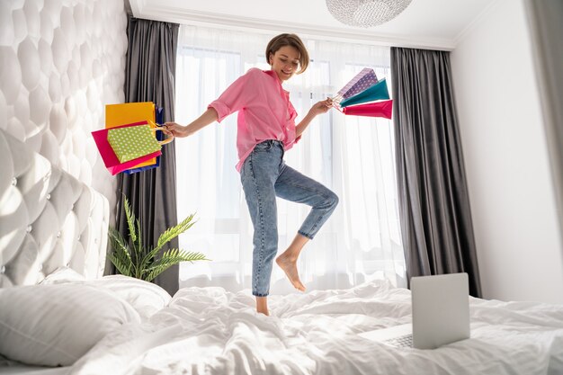 Donna abbastanza felice che si diverte a saltare sul letto a casa con borse della spesa colorate