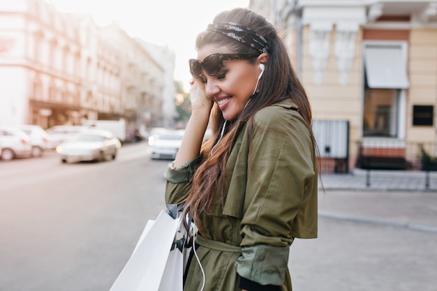 Donna abbastanza latina con nastro nero che ride per strada, ascoltando musica in auricolari