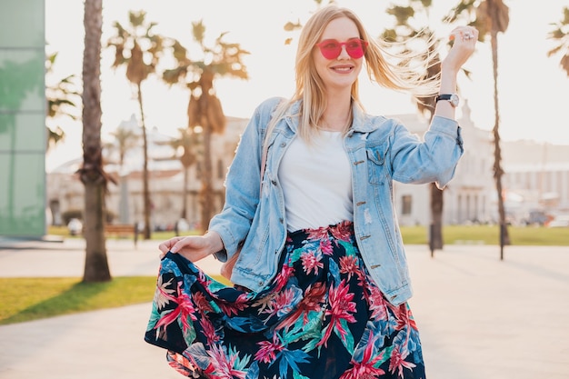 Donna abbastanza sorridente che cammina nella strada della città in elegante gonna stampata e giacca oversize in denim indossando occhiali da sole rosa, tendenza stile estivo