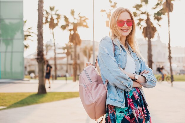 Donna abbastanza sorridente che cammina nella strada della città in elegante gonna stampata e giacca oversize in denim indossando occhiali da sole rosa, tendenza stile estivo