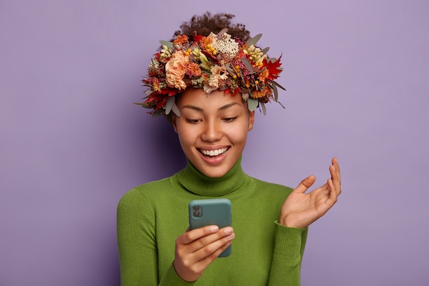 Donna abbastanza sorridente usa come un telefono cellulare e solleva il palmo e ridacchia positivamente, indossa una simbolica ghirlanda autunnale e un maglione verde