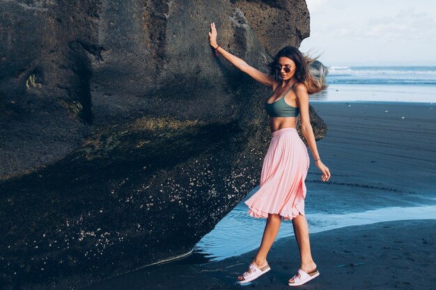 Donna abbronzata in forma caucasica in minuscolo top e gonna rosa sulla spiaggia di sabbia nera