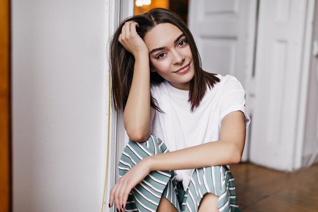 Donna accattivante leggermente abbronzata che posa a casa. Foto dell'interno dell'affascinante ragazza bruna seduta sul pavimento e tocca i suoi capelli.