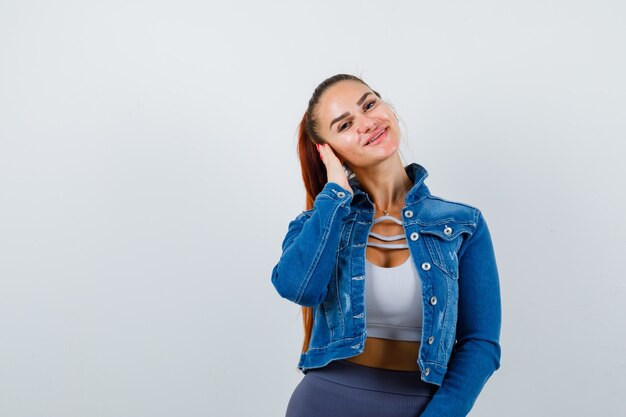 Donna adatta che preme la mano sull'orecchio, sorride in top corto, giacca di jeans, leggings e sembra felice, vista frontale.