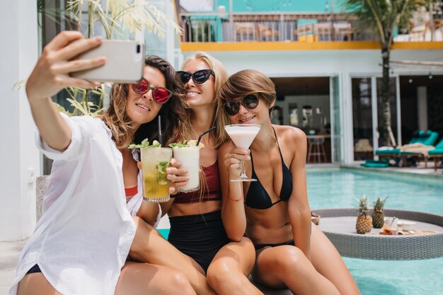 Donna adorabile con capelli scuri che tiene cocktail e che fa selfie. belle donne che si rilassano in piscina e posano per una foto.
