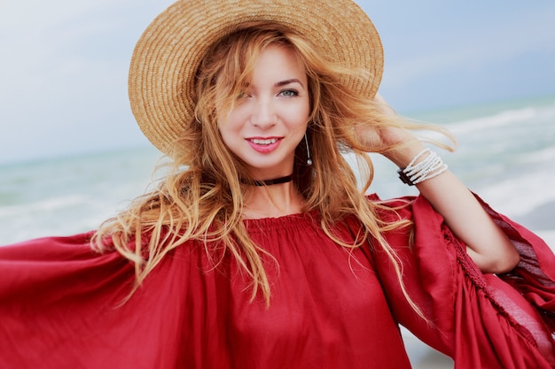 Donna adorabile in cappello di paglia e vestito rosso estivo alla moda in posa vicino alle onde dell'oceano. Sorriso schietto.