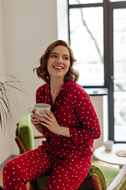 Donna adorabile in pigiama rosso che tiene tazza di caffè. Tiro al coperto di giovane donna sorridente che gode del tè a casa.