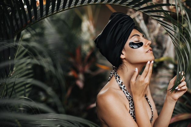 Donna adorabile in turbante nero in piedi sul fondo della natura. Colpo esterno della signora elegante con bende sugli occhi vicino alle palme.