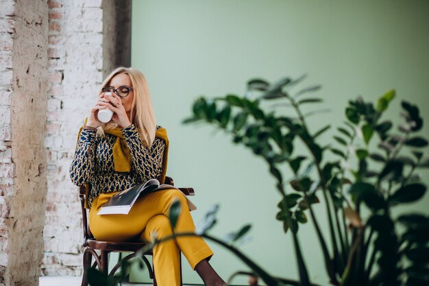 Donna adulta che si siede nella sedia che beve caffè e che legge rivista