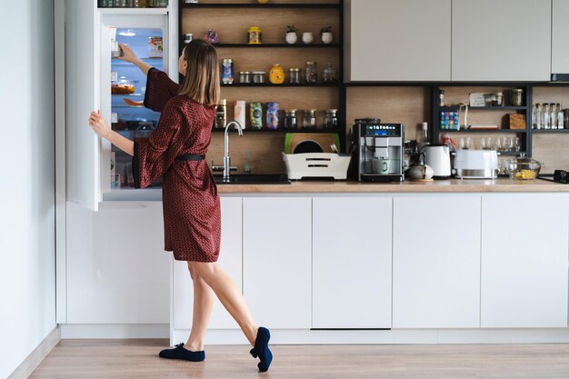 Donna affamata che cerca cibo nel frigorifero a casa ma non ha molto lì Mobili da cucina bianchi per la casa indossano un accappatoio di seta rossa
